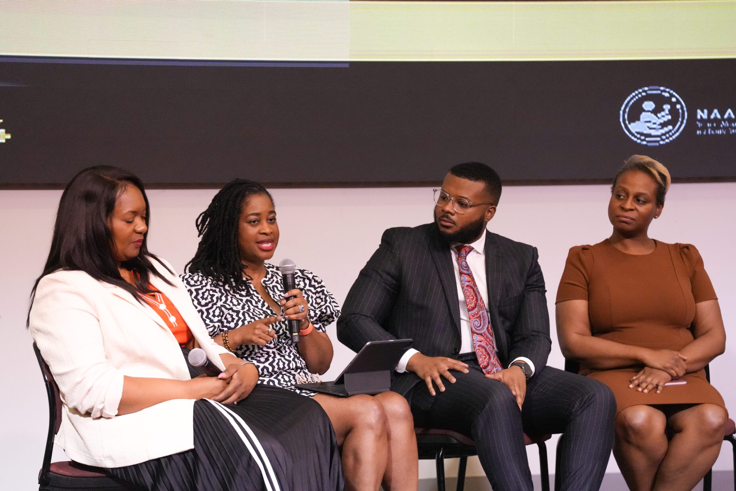 A panel of four people seated on stage at a conference. The woman on the left, wearing a white blazer, listens attentively. The woman next to her, wearing a patterned dress, is speaking into a microphone. The man next to her in a pinstripe suit and patterned tie looks on thoughtfully. The woman on the far right, wearing a brown dress, is also listening intently.